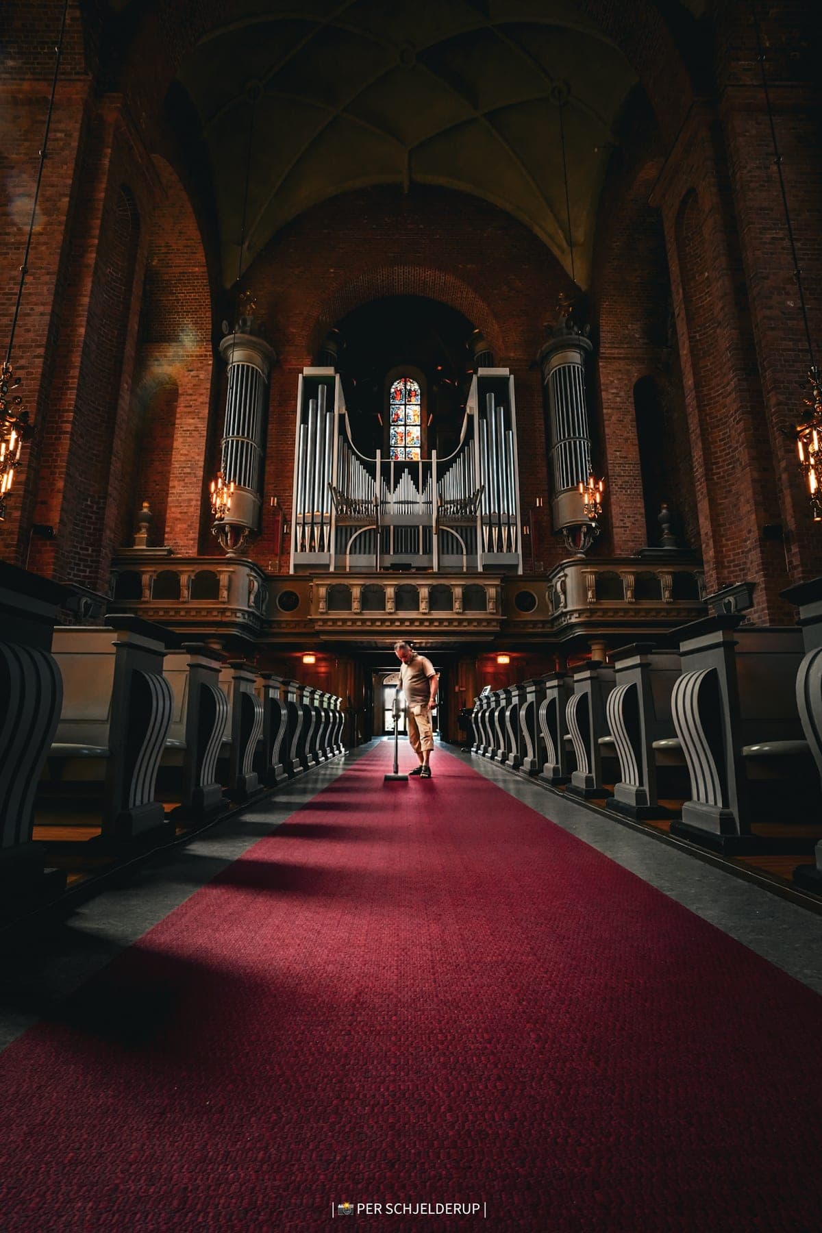 A red-carpeted church aisle leading towards the organ, a lone figure standing at the far end.